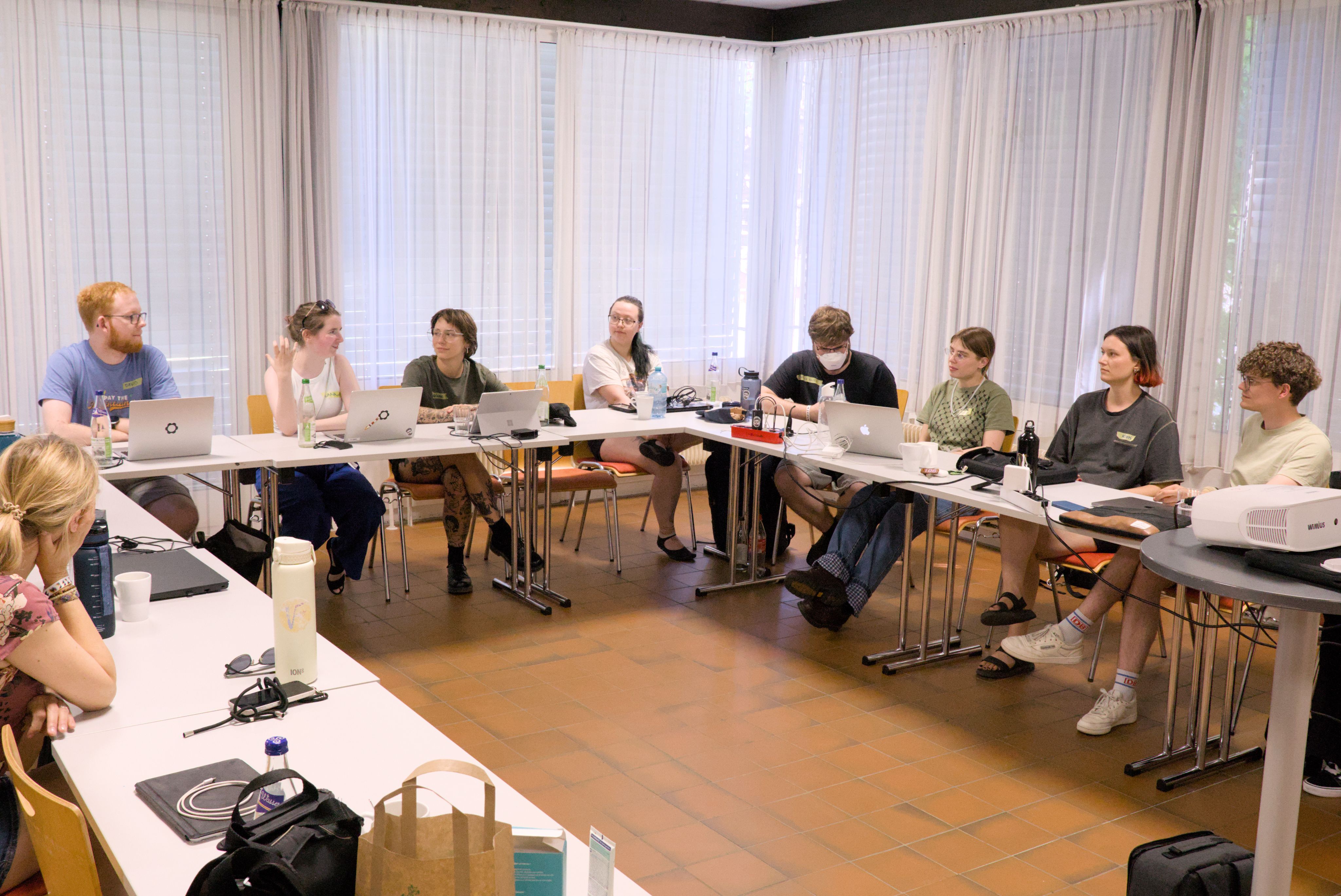 Viele unterschiedliche junge Leute sitzen um einen U-förmigen Workshoptisch herum und hören einer sprechenden Person zu.