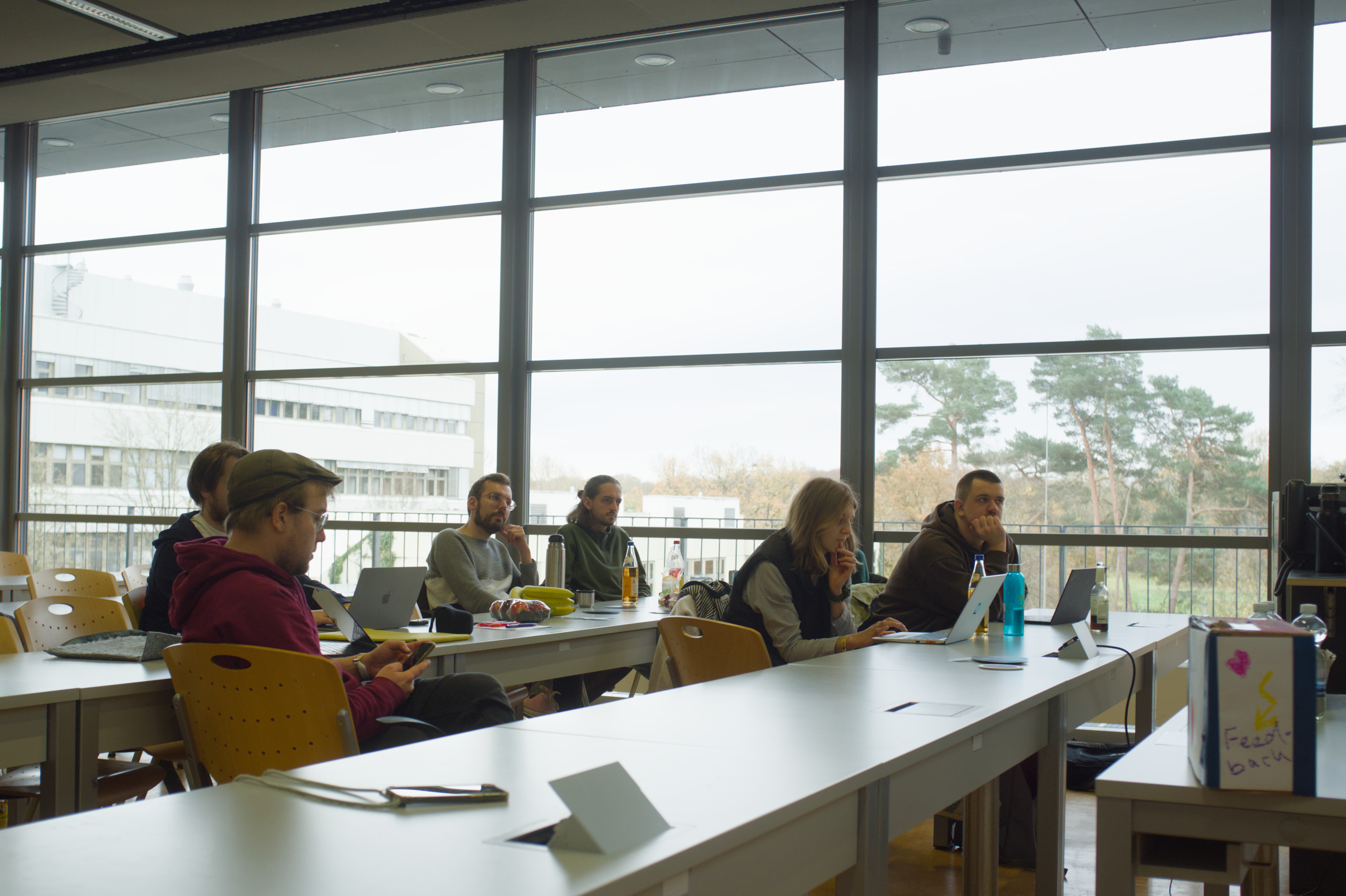 Blick von der Innenseite des Raums auf die große Fensterfront vor der die Personen sitzen und dem Vortrag von Lea lauschen.