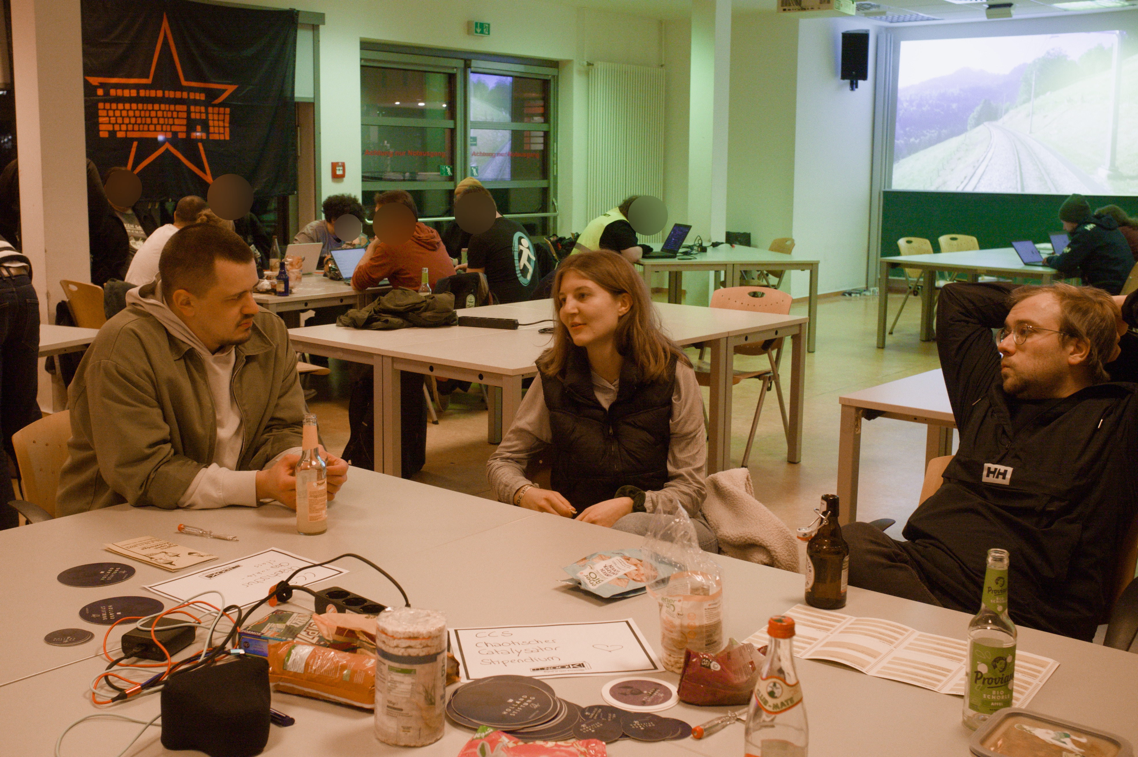 In einem bun beleuchteten Raum sitzen drei Personen an einem Tisch und unterhalten sich. Im Hintergrund ist sind Fahnen und LEDs, sowie weitere Tischgruppen zu sehen.